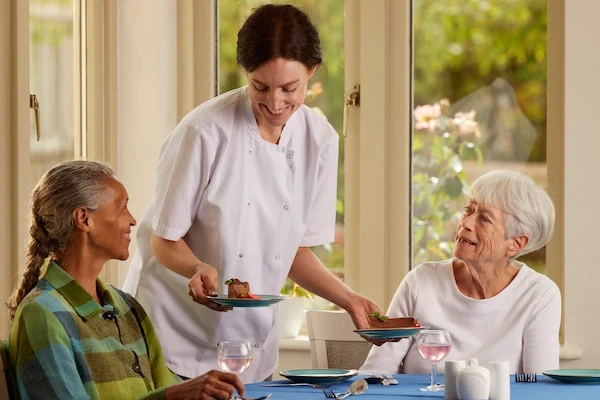 A carer serving tea to residents at a table by the window