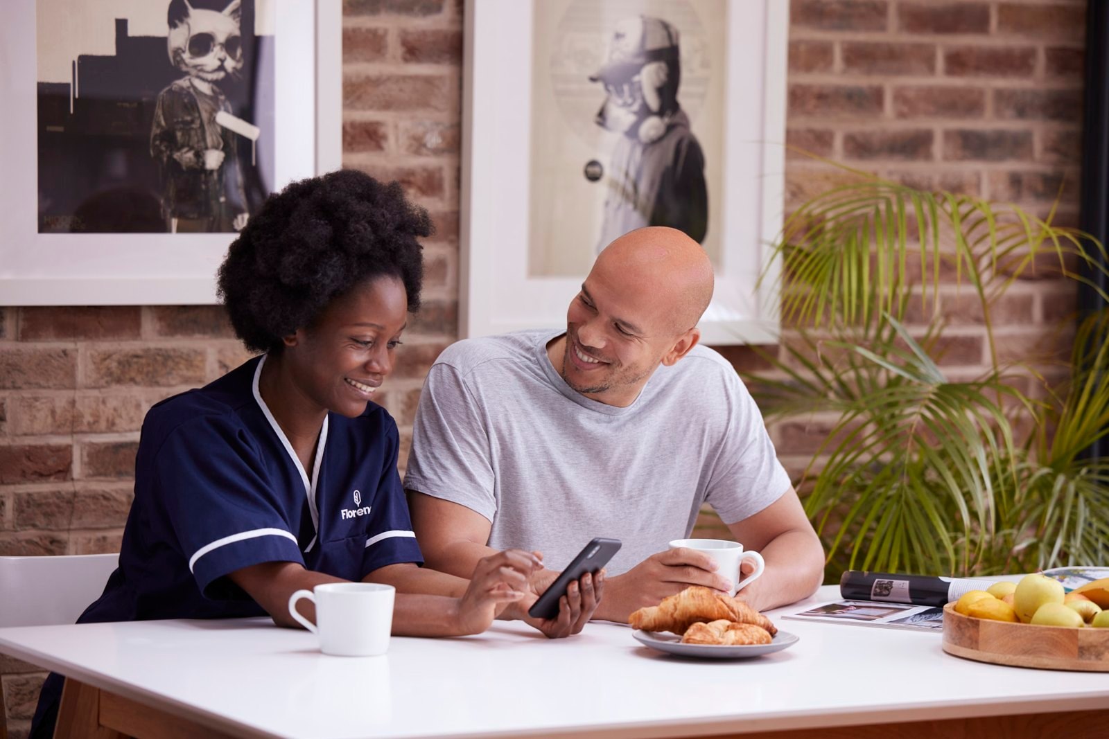 A nurse and a client laughing over a phone at a kitchen table