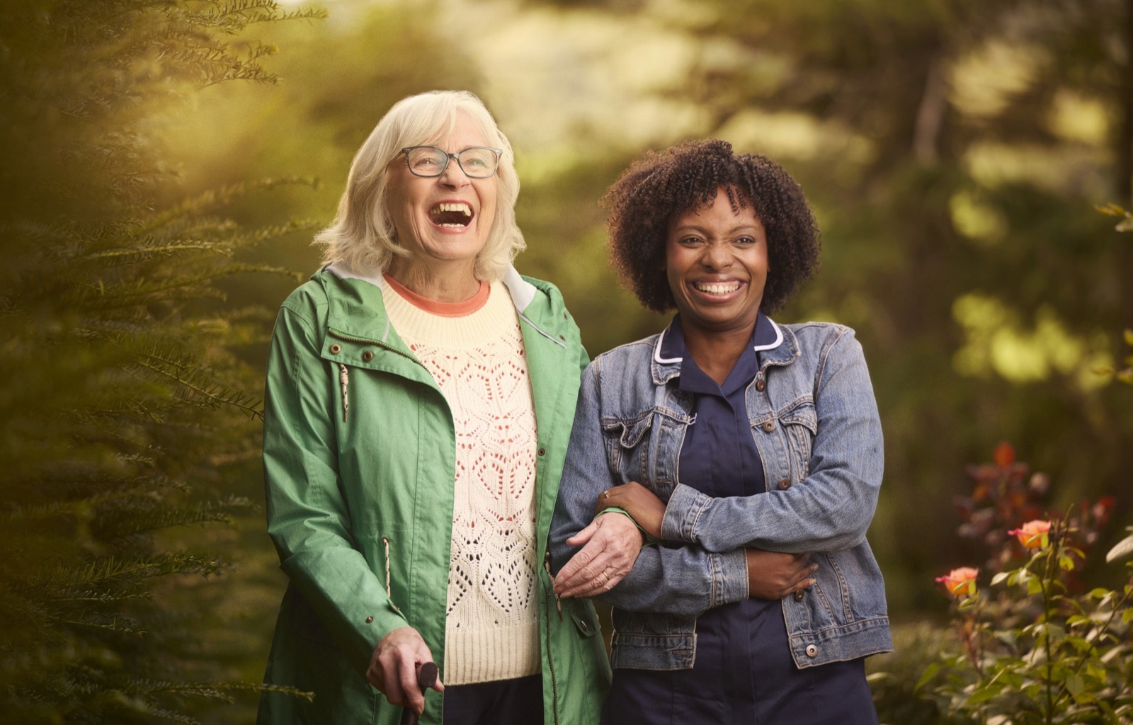 A carer and an older woman laughing arm in arm in a garden