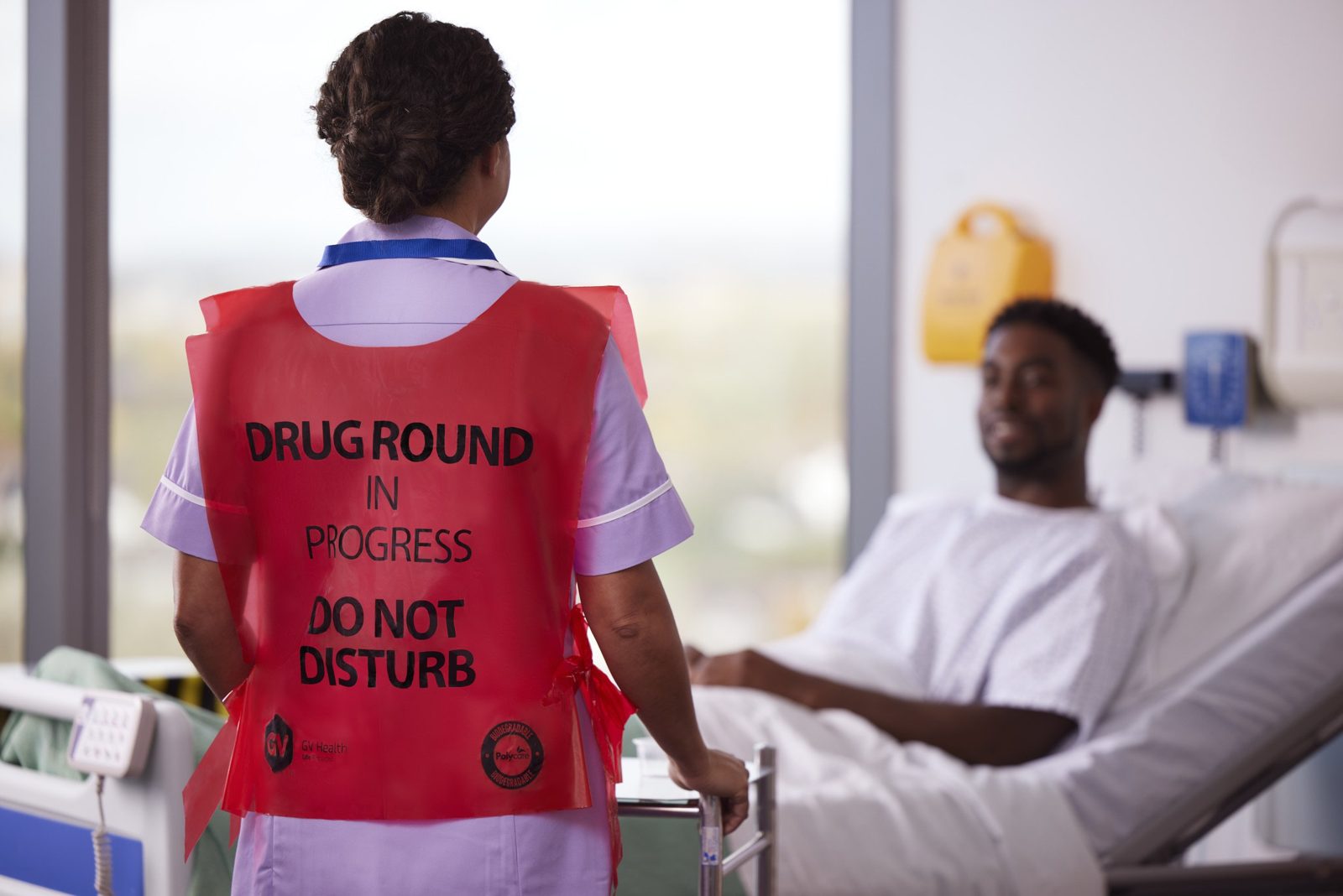 A nurse in a GV Health drug round tabard walking away from a patient smiling in bed